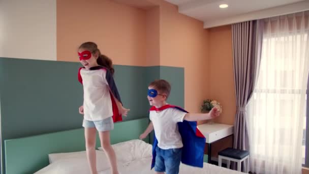 Lány és fiú szuperhősök, ugrálnak a szobában az ágyon, a gyerekszobában, két gyerek piros és kék Superman jelmezben, szuperhősök, testvér és nővér, játszanak otthon képzelve, hogy ők szuperhősök. — Stock videók
