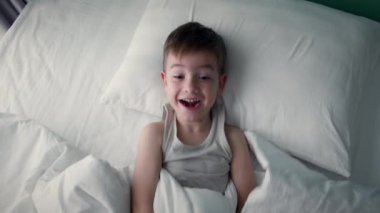 Mutlu, neşeli, beyaz pijamalı bir çocuğun portresi. Sabah yatağında uyanır, ailesine gülümserken bakar. Kahkahalar atar ve tekrar battaniyenin altına saklanır..