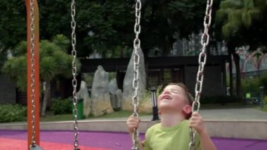 Mutlu gülen çocuk parkta salıncakta sallanıyor, güneşli bir günde salıncakta sallanıyor, neşeli bir çocuk yukarı aşağı uçuyor, çocuk parkta oynarken gülüyor ve gülümsüyor.,