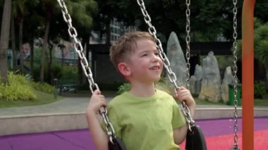 Mutlu gülen çocuk parkta salıncakta sallanıyor, güneşli bir günde salıncakta sallanıyor, neşeli bir çocuk yukarı aşağı uçuyor, çocuk parkta oynarken gülüyor ve gülümsüyor.,
