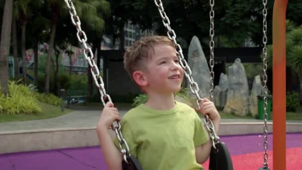 Glada skrattande barn gungar på en gunga i parken liten pojke, på en gunga i lekplatsen på en solig dag, ett glatt barn flyger upp och ner, barn skrattar och ler när du spelar i stadsparken, — Stockvideo