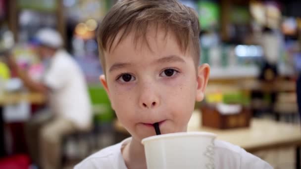 Kind tegen de achtergrond van bezoekers in een café kijkt naar de camera en drinkt sap. Een kind voeden in een café. Zoon jongen zit aan tafel koolzuurhoudend sap te drinken — Stockvideo