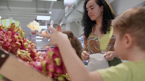 小さな幸せな息子と娘を持つかわいい女性は、食料品店で一緒に果物新鮮なドラゴンフルーツ、マンゴーを選択します。子供とお母さんは熟したレタスを取る棚から新鮮な緑の葉バスケットに入れ — ストック動画