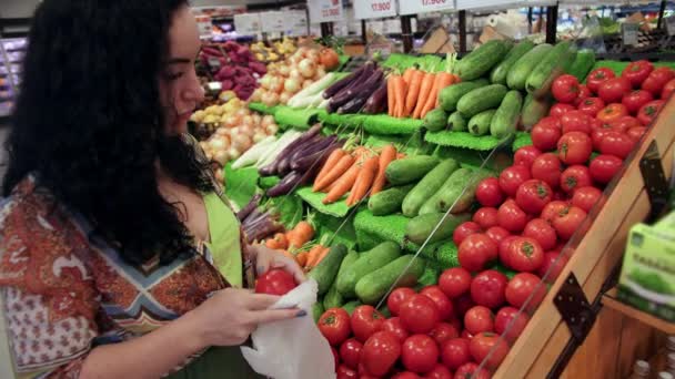 Mulher jovem dona de casa em uma mercearia compra leva tomates maduros da prateleira e coloca-los em um cesto., uma jovem escolhe no departamento de supermercado. — Vídeo de Stock