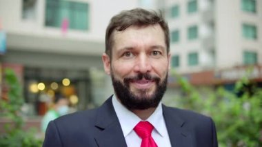 Yakışıklı iş adamı modern ticari kompleksin dışındaki kameraya bakıp gülümsüyor. Portre. İyi giyimli, mutlu sakallı ofis yöneticisi. Modern şirket binasının önünde duran şirket yöneticisi..