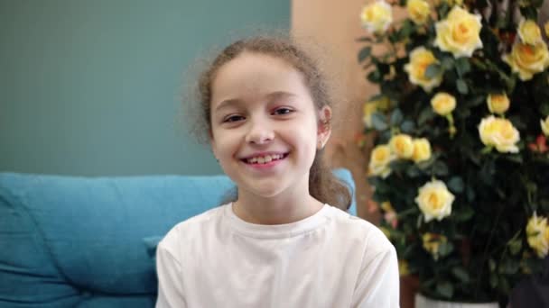 Ritratto bambino carino con bel viso seduto sul divano. Divertente bambina sorridente bambino guardando la fotocamera sono seduti sul divano a casa. Concetto di infanzia felice. — Video Stock