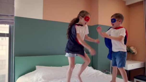 Супергерои, брат и сестра, играют дома, воображая себя супергероями. Девушка и мальчик супергерои, прыгают в комнате на кровати, в детской комнате, двое детей в красно-синем костюме Супермена, — стоковое видео