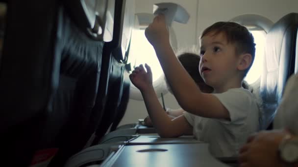 飛行機の窓のそばに座って、飛行機の飛行中に小さな紙飛行機で遊んでいる子供。飛行機の窓の後ろに小さな紙飛行機と子供の手. — ストック動画