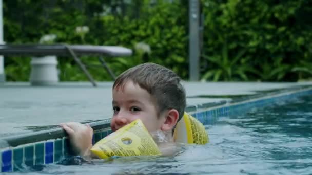 Маленький мальчик в бассейне с надувными браслетами на руках держится за край бассейна руками. Ребенок веселится и играет в бассейне. Летние каникулы, каникулы, рыдания — стоковое видео