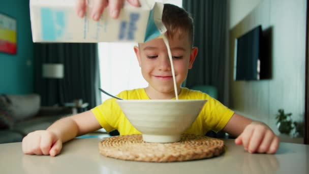 Ojciec nalewa mleko na płatki, przygotowuje śniadanie dla syna. Mały głodny uroczy chłopiec siedzi przy stole, je płatki kukurydziane z mlekiem z apetytem przyjemności, cieszy się śniadaniem. — Wideo stockowe