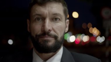 Pahalı takım elbiseli, yakışıklı, sakallı kıdemli bir işadamı sokakta binaların yanında dikiliyor ve kameraya gülümsüyor..