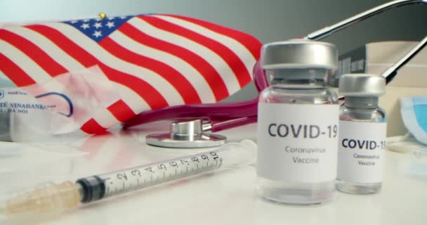 COVID-19 Impfstoff entwickelt, Coronavirus-Impfampulle und verschiedene Spritzen, medizinische Werkzeuge, Gesichtsschutz, Hintergrund der amerikanischen Flagge und Bücher. — Stockvideo