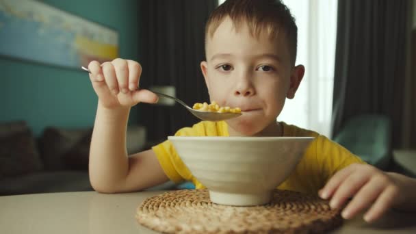 Πορτρέτο λίγο πεινασμένο παιδί χαριτωμένο αγόρι κάθεται στο τραπέζι, τρώει κορν φλέικς με γάλα με όρεξη της ευχαρίστησης, απολαμβάνει το πρωινό. — Αρχείο Βίντεο