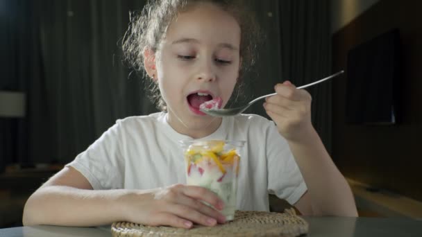 Дитина їсть молочний йогурт з тропічними фруктами. Здорове харчування маленької дівчинки обід концепція дитячої мрії. Голодна дочка дівчина на кухні їсть йогуртове молоко з фруктами. Позитивна дитина посміхається — стокове відео