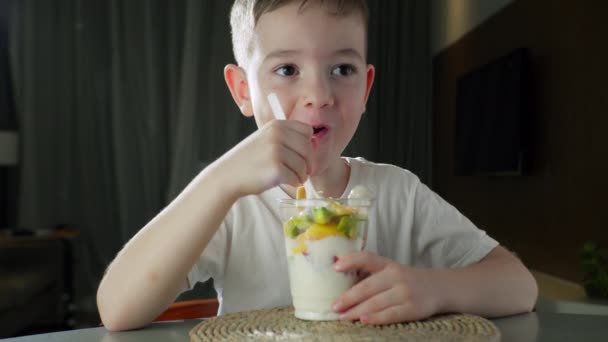 Child eats milk yogurt with tropical fruits. Healthy eating little boy dinner concept of childhood dream. Hungry daughter girl in the kitchen eats yogurt milk with fruits. Positive child smiling — Stock Video