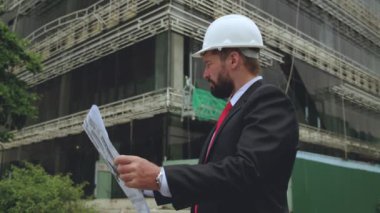 Sakallı, kıdemli mimar, müteahhit, beyaz kasklı, takım elbiseli, elinde bir proje, bir gökdelen ve bir arka plan tutan profesyonel Kafkas mühendisi portresi.