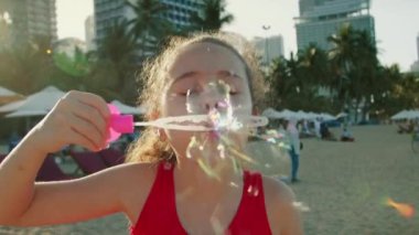 Komik, mutlu küçük kız gülümseyen çocuk kameraya bakıyor ve kumsalda oynuyor. Sabun köpüğüyle bebek oyunu. Gülümseyen mutlu çocuk gülümsemesi. Bebek gülüşü ve sabun köpükleri.
