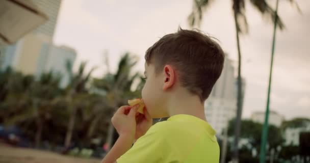 相貌饥饿有趣的小男孩心甘情愿地远眺一边吃着美味的披萨 一个可爱的孩子在海滩上远眺一边吃着意大利披萨 漂亮脸蛋的可爱孩子贪婪地吃着 快乐的童年4K — 图库视频影像