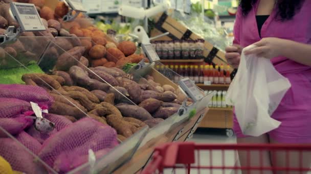 若い女性は食料品を購入し スーパーマーケットで体重計のために袋にジャガイモを拾います ピンクのスーツの魅力的な若い女性は果物や野菜市場でOを選択します — ストック動画