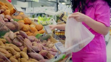Genç bir kadın market alışverişi yapıyor, süpermarkette tartılmak üzere patates topluyor. Pembe takım elbiseli çekici genç bir kadın meyve ve sebze pazarını seçiyor.