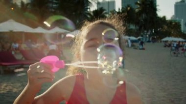 Komik, mutlu küçük kız gülümseyen çocuk kameraya bakıyor ve kumsalda oynuyor. Sabun köpüğüyle bebek oyunu. Gülümseyen mutlu çocuk gülümsemesi. Bebek gülüşü ve sabun köpükleri.