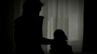 Ev karantinasında sıkılmış bir çocuk. Koruyucu tıbbi maskeli bir bebeği olan küçük bir kız yatakta oturur ve pencereden dışarı bakar. Alerji, covid-19 koronavirüs salgını, salgını önleme
