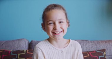 Evdeki kanepede kameraya bakıp gülümseyen komik küçük sevimli kız portresi, güzel yüzlü şirin çocuk kanepede oturuyor. Mutlu bir çocukluk kavramı