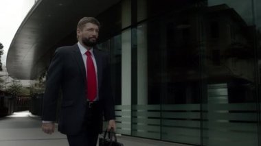 Pahalı takım elbiseli bir işadamı elinde evrak çantasıyla güneşli yaz gününde şehir caddesinde ağır çekimde yürüyor. Kavram işadamı, teknoloji, iletişim, başarı