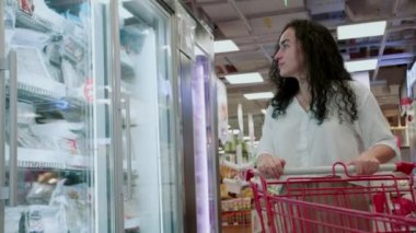 Beyaz takım elbiseli güzel bir kadın markette dondurulmuş prozhukt seçiyor marketteki taze deniz ürünleri. Genç bir kadın markette taze donmuş ürünleri seçiyor ve onları sepete koyuyor.