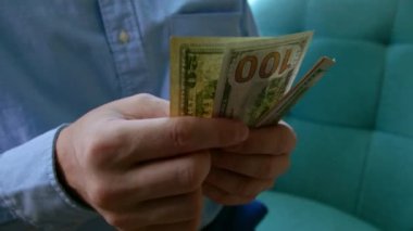 Adam gömleğinin cebinden 100 ve 50 dolarlık banknotlar veriyor, parayı sayıyor, sonra ikiye katlıyor ve gömleğinin cebine koyuyor. Mali yetersizlik, ellerinde dolar banknotları