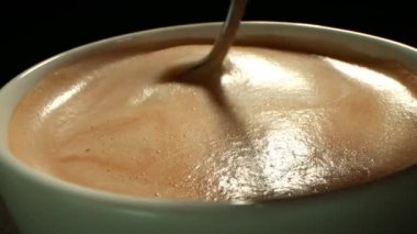 Kahveli bir fincanın üstünden köpüklü köpüğü karıştıran, kahveyi kaşıkla karıştıran birinin eli.