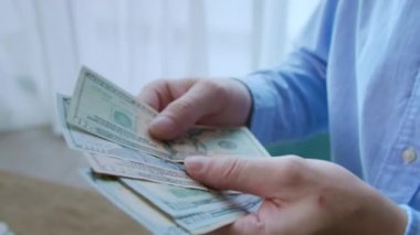 Adam gömleğinin cebinden 100 ve 50 dolarlık banknotlar veriyor, parayı sayıyor, sonra ikiye katlıyor ve gömleğinin cebine koyuyor. Mali yetersizlik, ellerinde dolar banknotları