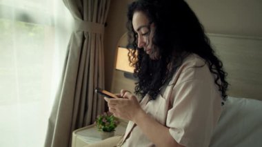Yatak odasında otururken akıllı telefon kullanan neşeli, genç Hintli kadın. Evde cep telefonu uygulaması kullanan akıllı, gülümseyen bir kadın. Güzel bir kız cep telefonuyla konuşurken rahatlıyor ve telefona bakıyor.