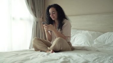 Yatak odasında otururken akıllı telefon kullanan neşeli, genç Hintli kadın. Evde cep telefonu uygulaması kullanan akıllı, gülümseyen bir kadın. Güzel bir kız cep telefonuyla konuşurken rahatlıyor ve telefona bakıyor.
