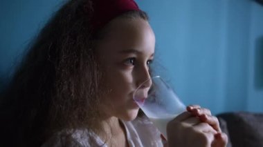 Küçük kız süt içiyor. Çocuk kahvaltısı yiyerek sağlıklı beslenme hayali. Oturma odasındaki kanepede oturan kız yoğurtlu süt içiyor ve kefir dudaklarını yalıyor. pozitif çocuk gülümsüyor