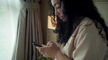 Yatak odasında otururken akıllı telefon kullanan neşeli, genç Hintli kadın. Evde cep telefonu uygulaması kullanan akıllı, gülümseyen bir kadın. Güzel bir kız cep telefonuyla konuşurken rahatlıyor ve telefona bakıyor.