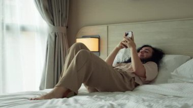 Güzel bir kız cep telefonuyla sohbet ederken ve telefona bakarken dinleniyor. Yatak odasında otururken akıllı telefon kullanan beyaz bir kadın. Evdeki cep telefonunda uygulamayla gülümseyen bir kadın.