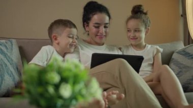 Mutlu aile anne ve şirin küçük çocuklar gülüyor akıllı tablet komik yüz maskesi uygulaması hücre ekranına bak, çocuklu anne cep telefonuyla video çağrısı yapıyor evdeki koltukta otur.