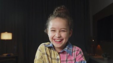 Portre Komik küçük kız kameraya bakıyor evde kanepede oturuyor, güzel yüzlü şirin çocuk kanepede oturuyor. Mutlu bir çocukluk kavramı. 4K.