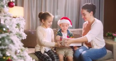 Noel Baba şapkalı küçük kızı ve oğlu olan mutlu aile Noel sabahı Noel ağacının yanında toplandı ve hediyelerin açılışının keyfini çıkardılar. Yeni yıl kutlaması, hediye mağazası reklam konsepti.