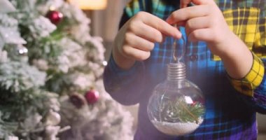 Çocuk elinde Noel ağacı için çok güzel bir Noel balosu tutuyor. Mutlu küçük çocuk Noel ağacını toplarla süslüyor. Ağacın altında bir sürü hediye kutusu var. Çocuk Noel ağacını süslüyor