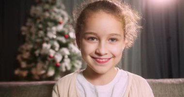 Noel şenliğinde portre çocuğu, evdeki koltukta oturmuş gülümseyen küçük kız arka plandaki dekoratif Noel ağacına bakıyor.