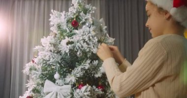 Çocuk elinde Noel ağacı için çok güzel bir Noel balosu tutuyor. Mutlu küçük kız Noel ağacını toplarla süslüyor. Ağacın altında bir sürü hediye kutusu var. Çocuk Noel ağacını süslüyor
