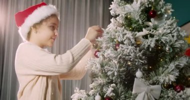 Çocuk elinde Noel ağacı için çok güzel bir Noel balosu tutuyor. Mutlu küçük çocuk Noel ağacını toplarla süslüyor. Ağacın altında bir sürü hediye kutusu var. Çocuk Noel ağacını süslüyor
