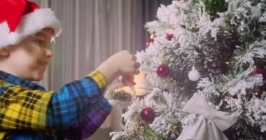Çocuk elinde Noel ağacı için çok güzel bir Noel balosu tutuyor. Mutlu küçük çocuk Noel ağacını toplarla süslüyor. Ağacın altında bir sürü hediye kutusu var. Çocuk Noel ağacını süslüyor