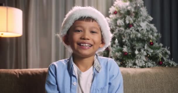 クリスマスの肖像画の子供 クリスマスの帽子の小さな男の子の笑顔サンタクロースはソファの上に座っている家でカメラを見て装飾的なクリスマスツリー — ストック動画