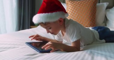 Noel Baba şapkalı mutlu, meraklı çocuk. Çocuk, yatak odasında tek başına duran dijital tablet teknolojisi cihazını kullanıyor. Evde küçük çocuk tutucu bilgisayar sörfü internet oyun oyunu.