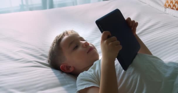 クリスマスの帽子の不思議な幸せなかわいい笑顔の子供サンタ クラウサ 単独で寝室に横たわっているデジタルタブレット技術デバイスを使用して子供の男の子 小さな子供は自宅でインターネットゲームをサーフィンパッドコンピュータを保持 — ストック動画