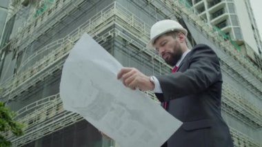 Güvenlik Üniforması ve Sert Şapka Giyen Profesyonel Kafkas Mühendis İşçi Portresi. Şema çizim planını kullanır. Kafkasyalı kendine güvenen endüstri uzmanı Fabrikada duruyor.