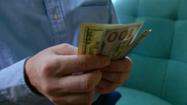 Adam gömleğinin cebinden 100 ve 50 dolarlık banknotlar veriyor, parayı sayıyor, sonra ikiye katlıyor ve gömleğinin cebine koyuyor. Mali yetersizlik, ellerinde dolar banknotları